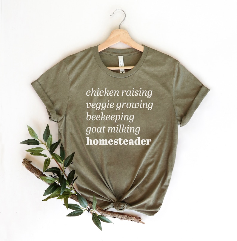 May include: A green t-shirt with white text that reads "chicken raising veggie growing beekeeping goat milking homesteader".