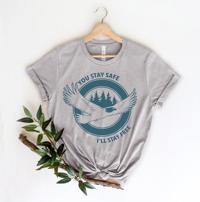 K&ouml;nnte beinhalten: Ein heidefarbenes T-Shirt mit einer blauen Grafik eines Adlers, der &uuml;ber einem Wald fliegt, mit dem Text "You Stay Safe I'll Stay Free".