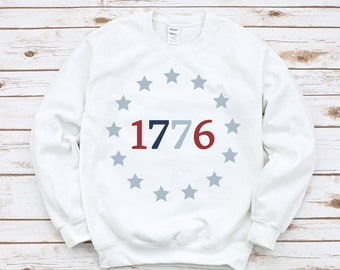 May include: White sweatshirt with a blue, red, and white graphic of the number 1776 surrounded by a circle of gray stars.