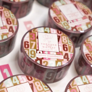 May include: Close-up of several rolls of decorative tape. Each roll features a repeating pattern of numbers in various colors, including pink, green, and brown. The tape is labeled "retro numbers" and includes the designer's name.