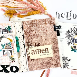 May include: A white and brown journal with the word "amen" and the text "a prayer journal" on the cover. The journal is decorated with black and white polka dots and floral designs. The journal is open to reveal a blank page with a black and white floral design and the word "XO" in a gold circle.