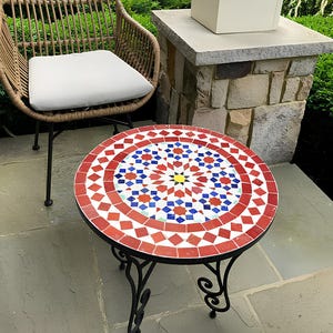 Puede incluir: Una mesa redonda de mosaico con un patrón geométrico rojo, azul y blanco. La mesa tiene una base de metal negro con un diseño de desplazamiento.
