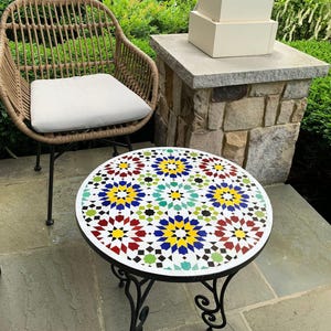 Puede incluir: Una mesa redonda de mosaico con un marco de metal negro. La superficie de la mesa presenta un patrón geométrico en tonos de rojo, amarillo, azul, verde y blanco. La mesa está situada en un patio.