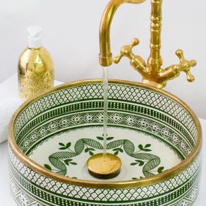 May include: A decorative ceramic sink with a green and white patterned design. The sink has a gold rim and a gold faucet with water flowing into it. A gold soap dispenser is visible in the background.