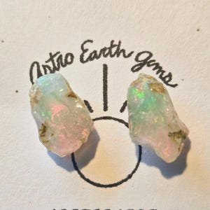 May include: Two raw opal gemstones with iridescent flashes of color. The opals are light in color with pink and green hues. The image includes the text "Astro Earth Gems" in a handwritten font.