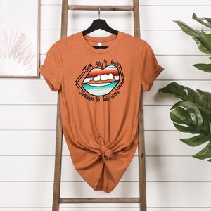 May include: A rust-colored t-shirt with a graphic of a mouth with a rainbow stripe design and the text "Then you'll have something to talk about".