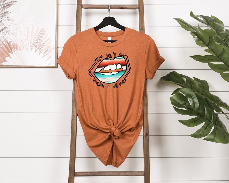 May include: A rust-colored t-shirt with a graphic of a mouth with a rainbow stripe design and the text "Then you'll have something to talk about".