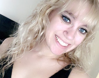 May include: A smiling person with blonde, wavy hair and blue eyes. They are wearing a black top. The background is a light-colored wall and ceiling. The person has a nose piercing.
