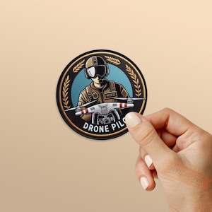 May include: A round patch featuring a drone pilot illustration. The design includes a pilot in a helmet and goggles, holding a drone. The text "DRONE PILOT" is displayed in a circular pattern. The patch has a black border with gold accents.