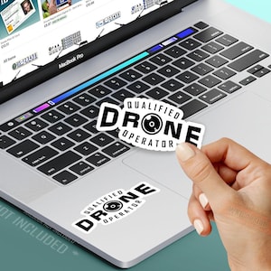 Könnte beinhalten: Ein weißer Aufkleber mit dem Text "QUALIFIED DRONE OPERATOR" in Schwarz wird über einer Laptop-Tastatur gehalten. Ein weiterer Aufkleber ist auf dem Laptop angebracht. Der Laptop ist silberfarben und hat eine schwarze Tastatur.