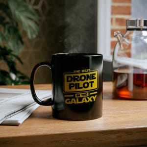 以下が含まれることがあります： 「The Best Drone Pilot In The Galaxy」という黄色のグラフィックが描かれた黒いコーヒーカップです。