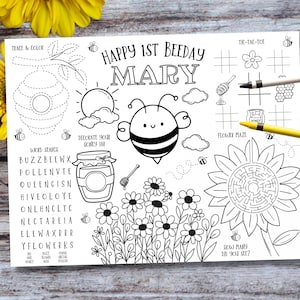May include: A black and white coloring sheet with a bee theme. The sheet includes a bee, flowers, a honeycomb, and the text "Happy 1st Beeday Mary". It also features a word search, a tic-tac-toe game, and a flower maze.