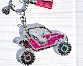 May include: A glittery keychain featuring a pink and silver off-road vehicle design. The keychain has a pink tassel and a small charm. The wheels and vehicle details are outlined in black.