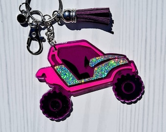May include: Pink and silver glitter keychain featuring a pink off-road vehicle with a black interior. The keychain has a silver tassel.