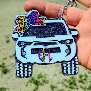 May include: A white glitter car charm with a black glitter background and a rainbow leopard print bow on top. The car has a black grill and headlights. The charm has a black and white tire tread design on the bottom.