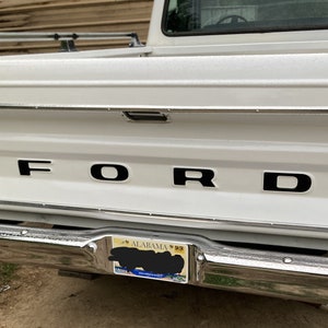 May include: A white Ford pickup truck with a black FORD logo on the tailgate. The truck has a chrome bumper and a license plate that reads "ALABAMA" with the number "0424".