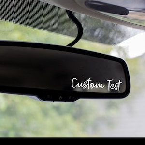 May include: A black rearview mirror with a white sticker that says "Custom Text" on it.