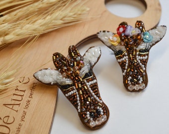 Peut inclure: Deux broches de girafe en perles avec des perles brunes, blanches et noires. Une broche a une couronne de perles colorées sur la tête. Les broches sont sur une surface en bois avec des épis de blé.