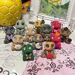 Puede incluir: Una colección de 11 pequeñas figuritas coloridas pintadas a mano. Las figuritas están hechas de arcilla polimérica y tienen una variedad de diseños caprichosos, incluyendo mariposas, flores y estrellas. Las figuritas están dispuestas sobre una superficie con estampado rosa y blanco.