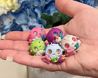 Puede incluir: Cinco pequeñas figuritas coloridas hechas a mano con caras caprichosas y detalles de purpurina. Cada figurita tiene forma de criatura marina y un diseño único.