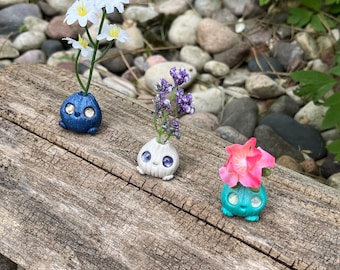 Puede incluir: Tres pequeños jarrones de cerámica con caras y ojos saltones, cada uno con un tipo diferente de flor. Los jarrones son azules, blancos y verdes. Las flores son blancas, moradas y rosas.