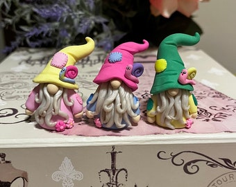 May include: Three small gnome figurines made of polymer clay. Each gnome has a different colored hat: yellow, pink, and green. Each gnome has a white beard and a snail on their hat.