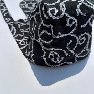 May include: A black head wrap or bandana featuring a repeating white and grey bicycle chain link pattern. The fabric is shaped with a rounded top section and two long ties extending from the sides, designed to be secured around the head.