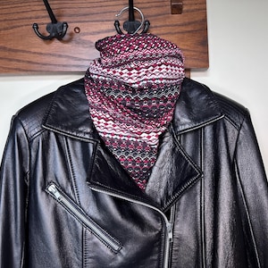 May include: A black leather jacket with a silver zip and a patterned scarf hanging on a wooden coat rack. The scarf has a geometric design in burgundy, white, and black.