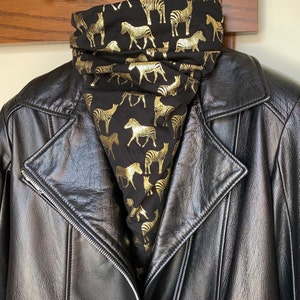 May include: A black leather jacket with a gold zebra print cowl neck warmer. The cowl neck warmer is black with a repeating pattern of gold zebras. The jacket has a zip closure and a notched collar.