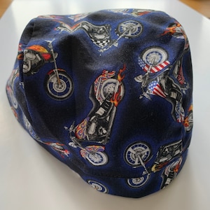 May include: A dark blue scrub cap featuring a repeating pattern of motorcycles with flames and American flag motifs. The head covering has a rounded crown and a stitched hem, displaying various motorcycle designs in shades of red, white, and black.