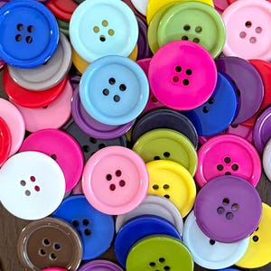 May include: A collection of round, four-hole plastic buttons in a vibrant assortment of colors including red, blue, yellow, green, pink, purple, white, brown, and gray. The buttons are scattered in an overlapping arrangement, showing various sizes.