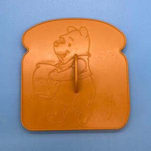 May include: Orange plastic Winnie the Pooh cookie cutter shaped like a slice of bread with Winnie the Pooh holding a pot of honey.