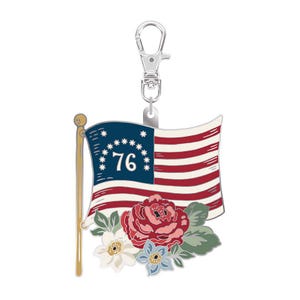 May include: A patriotic charm featuring a stylized American flag with red and white stripes, a blue field with stars, and the number "76". Below the flag are floral accents in shades of pink, white, and blue. The charm is attached to a silver-colored clasp.