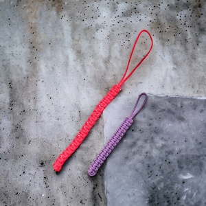 Op de afbeelding: Twee ritslipjes van paracord, één in felrood en één in paars. Elk lipje heeft een strak geweven, getextureerd knooppatroon en een flexibele lus aan de bovenkant voor bevestiging. Het rode lipje is langer dan het paarse lipje, beide van koord.