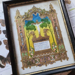 Può includere: Opera d'arte religiosa incorniciata raffigurante la crocifissione. L'opera d'arte ha una cornice dorata e ornata. L'immagine include una figura centrale sulla croce, angeli e testo in tedesco. Lo sfondo è giallo e blu.