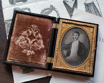 May include: An open, antique black case with a gold-framed portrait of a man in a suit. The left side of the case has a brown velvet lining with an embossed design. Vintage ephemera surrounds the case.