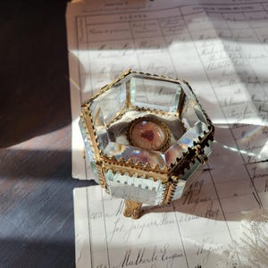 May include: A hexagonal glass jewellery box with bevelled edges and a gold-toned metal frame. The box features a hinged lid and stands on small decorative metal feet. Inside, a small circular medallion with a red heart illustration is visible.