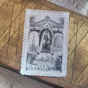 Op de afbeelding: Een rechthoekige witte papieren kaart met een decoratieve gestanste kanten rand, voorzien van een zwart-witte illustratie van een religieuze plek. De centrale afbeelding toont een figuur in gewaad met een kind, gelabeld Souvenir de Ste Anne d'Auray.