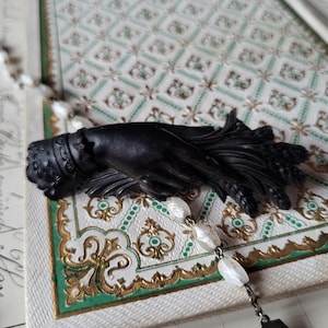 May include: A black, ornate hand sculpture with detailed fingers and wrist adornments. The sculpture rests on a decorative surface with a green and gold border, and is accompanied by a beaded chain with white, oval-shaped beads.