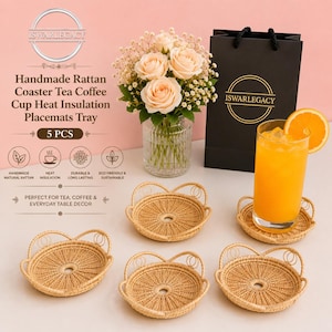 May include: A set of five round, light brown woven rattan coasters with small decorative loops along the edges. Each coaster features a central circular opening. The coasters are arranged on a surface alongside a glass of orange juice and a black gift bag.
