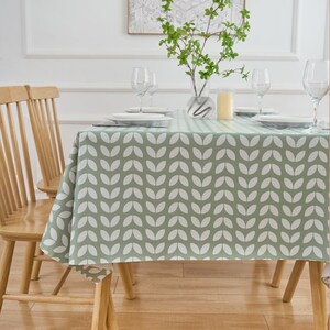 May include: A sage green tablecloth with a white leaf pattern covers a wooden table set for a meal. The table is set with white plates, wine glasses, and a vase of greenery. The wooden chairs have a light brown finish.