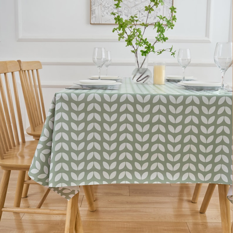 May include: A sage green tablecloth with a white leaf pattern covers a wooden table set for a meal. The table is set with white plates, wine glasses, and a vase of greenery. The wooden chairs have a light brown finish.