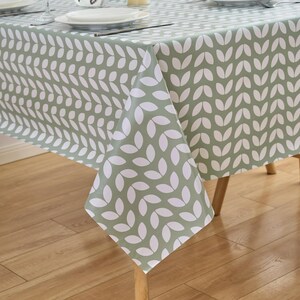 May include: A sage green tablecloth with a repeating white leaf pattern. The tablecloth is draped over a wooden table, with a portion of the table setting visible. The leaf design is arranged in vertical rows, creating a textured appearance.