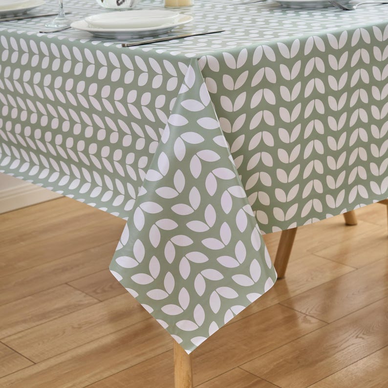 May include: A sage green tablecloth with a repeating white leaf pattern. The tablecloth is draped over a wooden table, with a portion of the table setting visible. The leaf design is arranged in vertical rows, creating a textured appearance.