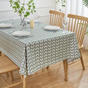 May include: A rectangular tablecloth with a repeating white leaf pattern on a sage green background. The table is set with white plates, wine glasses, and silverware. A vase of greenery and a candle are also on the table.