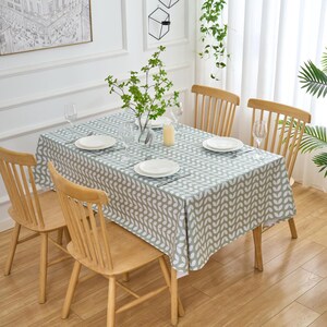May include: A dining table set for four, featuring a light green tablecloth with a white leaf pattern. The table is surrounded by four wooden chairs. The scene is set in a well-lit room with a painting and decorative plants.
