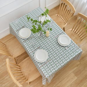 May include: Overhead view of a dining table set for a meal. The table is covered with a green and white leaf-patterned tablecloth. White plates, silverware, and glasses are set for each place setting. Wooden chairs surround the table.