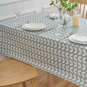 May include: A sage green tablecloth with a repeating white leaf pattern. The table is set with white plates, silverware, and wine glasses. A wooden chair is in the foreground, and a vase with greenery and a candle are on the table.