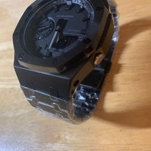 May include: A black G-Shock watch with a digital display and a black metal band. The watch has a square face with a black bezel and a black dial. The band is made of metal and has a black finish.