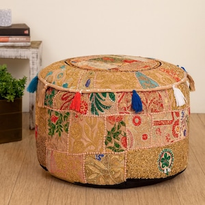 May include: A circular patchwork ottoman with a beige base and vibrant embroidered patches. It features tassels in blue, red, and white. The base is black, and the top has a circular design. The ottoman's diameter is approximately 60 cm.